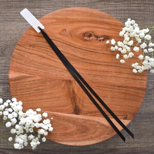 May include: A round, wooden cutting board with a natural wood grain pattern. Two black ribbons are laid across the board, with a white tag attached to one end. Sprigs of white baby's breath flowers are arranged around the board on a dark wood surface.