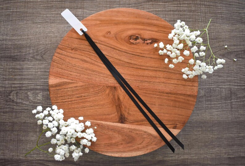 May include: A round, wooden cutting board with a natural wood grain pattern. Two black ribbons are laid across the board, with a white tag attached to one end. Sprigs of white baby's breath flowers are arranged around the board on a dark wood surface.