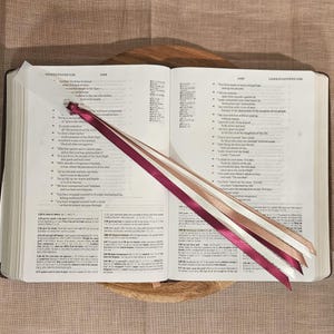 May include: An open book with text, featuring three colorful ribbon bookmarks. The ribbons are burgundy, cream, and light pink. The book is on a wooden tray, set against a neutral-toned fabric background.