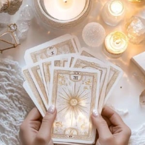 May include: A deck of tarot cards with gold and cream-colored designs, held in hands. The cards feature sunburst and geometric patterns. Lit candles and crystals surround the cards, creating a mystical ambiance.