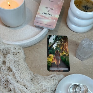 Peut inclure: Un jeu de cartes oracle Astro & Timing de Clémence et Philippe Lhoste, une carte illustrant une personne marchant sur un chemin, une bougie blanche allumée, une tasse blanche avec du café, un cristal transparent et des créoles en argent.