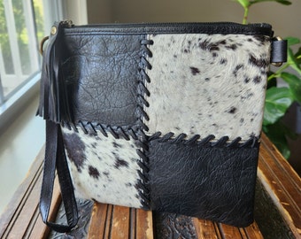 May include: A patchwork-style handbag featuring a combination of black leather and cowhide with black and white spots. The bag has a zipper closure, a wrist strap, and a decorative tassel. The stitching is visible and adds to the rustic aesthetic.