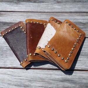 May include: Four leather wallets with cowhide accents in shades of brown, tan, and white. Each wallet is rectangular with rounded corners and a border of brass studs. The wallets are arranged on a weathered wooden surface.