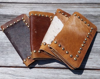 May include: Four leather wallets with cowhide accents in shades of brown, tan, and white. Each wallet is rectangular with rounded corners and a border of brass studs. The wallets are arranged on a weathered wooden surface.