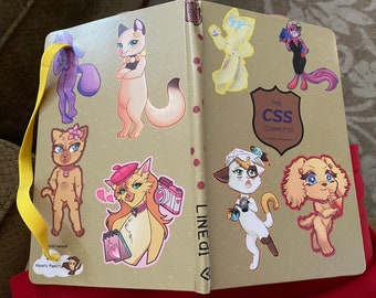 May include: A gold notebook with a yellow strap and various colorful cat stickers. The stickers feature cats in different poses and outfits, including a cat wearing a beret and holding a camera, a cat wearing a flower and a collar, and a cat wearing a black dress and holding a gun. The notebook also has a sticker with the text "The CSS Committee".