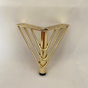 May include: Gold metal furniture leg with a triangular base and four horizontal bars. The leg has a black plastic foot.