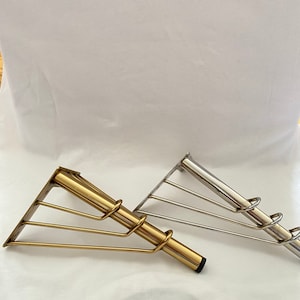 May include: Two metal shelf brackets, one gold and one silver, with a triangular base and a curved support bar. The brackets are designed to support shelves.