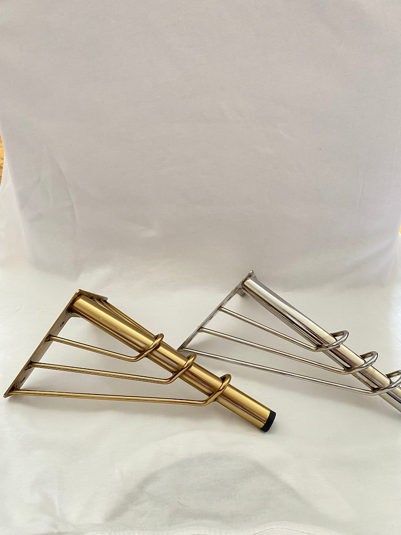 May include: Two metal shelf brackets, one gold and one silver, with a triangular base and a curved support bar. The brackets are designed to support shelves.