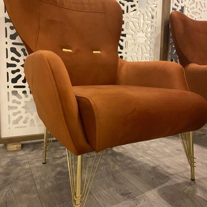 May include: A burnt orange velvet armchair with a high back and a unique gold metal base with multiple legs. The chair is sitting on a wood floor.