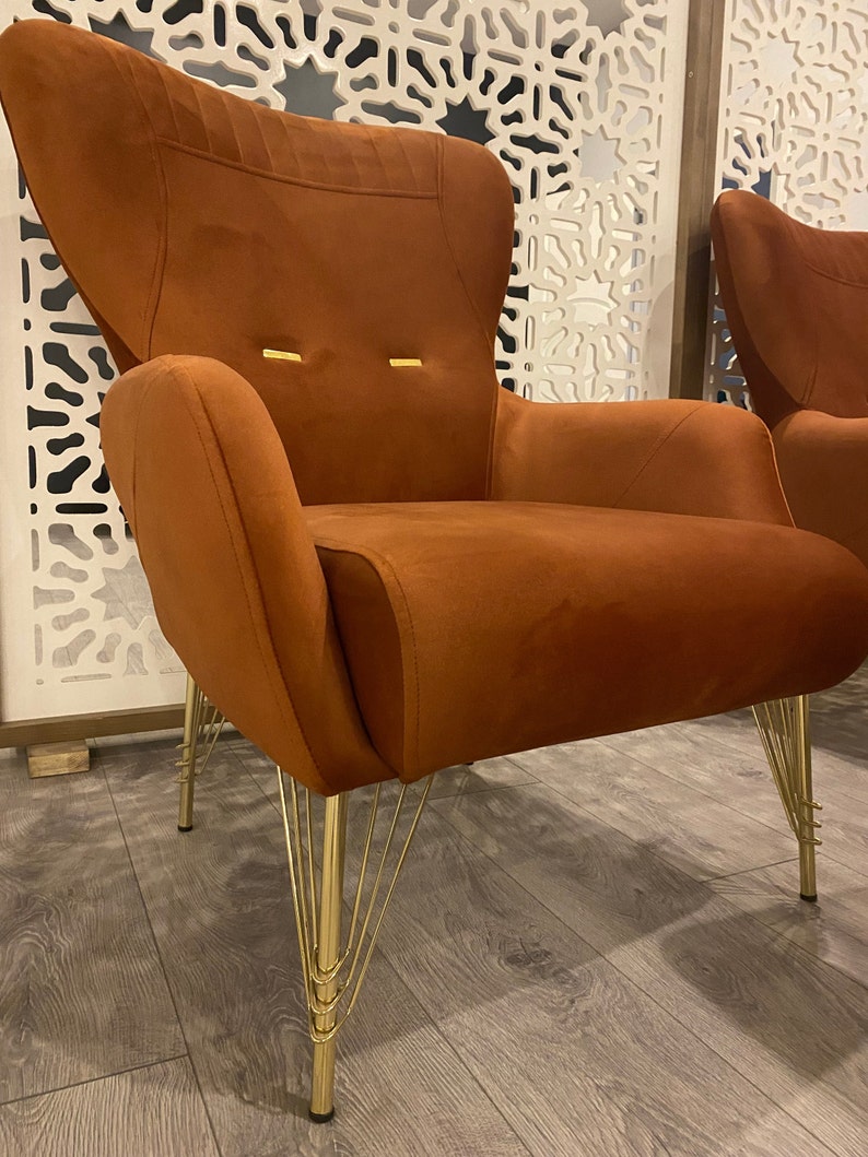 May include: A burnt orange velvet armchair with a high back and a unique gold metal base with multiple legs. The chair is sitting on a wood floor.