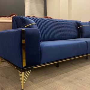 May include: A blue velvet sofa with gold trim and black legs. The sofa has a tufted back and is sitting on a wooden floor.
