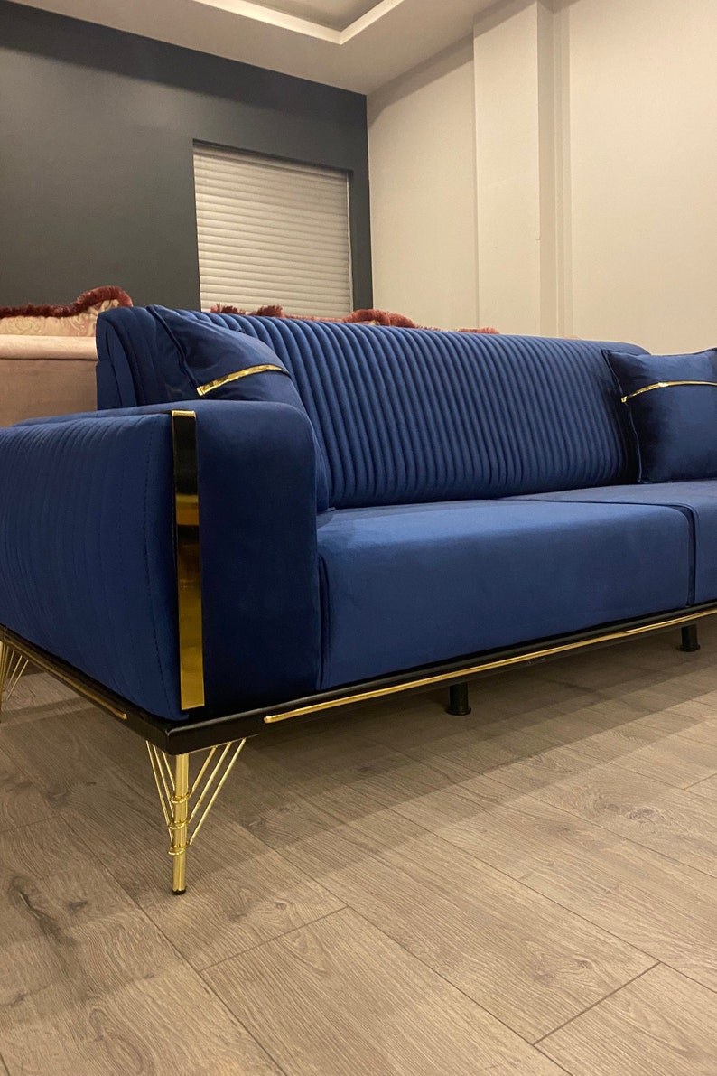 May include: A blue velvet sofa with gold trim and black legs. The sofa has a tufted back and is sitting on a wooden floor.