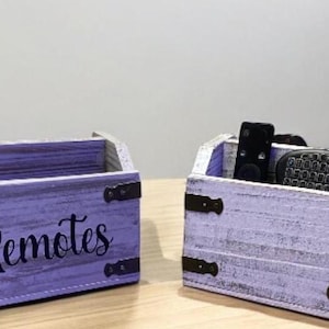May include: Two rustic, lavender-colored wooden remote control organizers. The boxes have a distressed finish, metal corner accents, and the word "Remotes" in black script. One box contains remotes.
