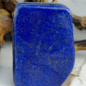 May include: A polished, rectangular lapis lazuli stone. The deep blue stone has gold flecks and a smooth surface. The stone is a rich, saturated blue colour. The edges are slightly rounded. The stone is a natural mineral.