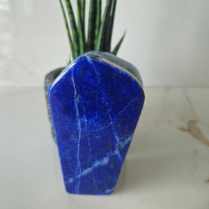 Puede incluir: Un cristal de lapislázuli azul oscuro, facetado, pulido con vetas blancas. El cristal tiene la forma de un rectángulo alto y estrecho.