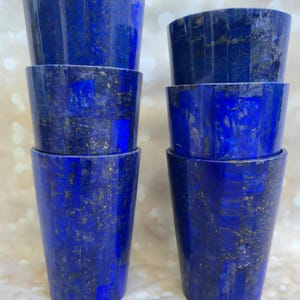 May include: Six blue lapis lazuli tumblers are stacked in two columns. The tumblers have a tapered shape and are a deep blue colour with gold flecks throughout the stone. The tumblers are arranged on a light-coloured surface.