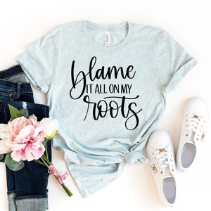 May include: A light blue t-shirt with the text "blame it all on my roots" in black script font.