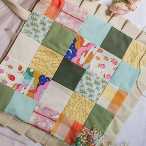 以下が含まれることがあります： 緑、青、黄、ピンク、オレンジなど、さまざまな色とパターンが組み合わされたパッチワークのデザインが施されたベージュのトートバッグ。バッグはフリルのある縁と茶色のストラップが付いています。