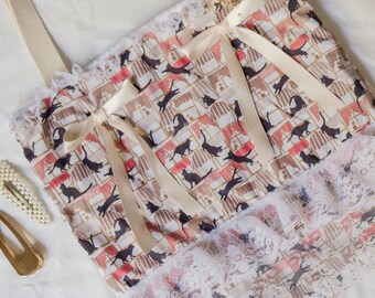 Puede incluir: Una bolsa de mano blanca con estampado de gatos blanco y negro y ribete de encaje. La bolsa tiene una correa de color crema y un lazo de color crema en la parte delantera.