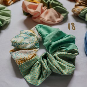 May include: A green and gold patterned scrunchie with a white background. The scrunchie is made of fabric and has a gathered look. The number 18 is written in brown on a white background.