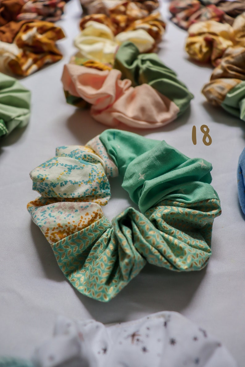 May include: A green and gold patterned scrunchie with a white background. The scrunchie is made of fabric and has a gathered look. The number 18 is written in brown on a white background.