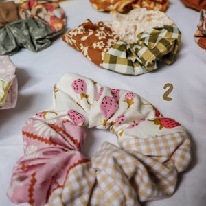 May include: A set of colorful fabric scrunchies with various patterns, including floral, gingham, and strawberry prints. The scrunchies are made of soft, stretchy fabric and are perfect for holding hair in place.