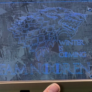 Op de afbeelding: Een rechthoekig, transparant acryl bord met een blauw geëtst ontwerp. Het ontwerp toont een wolfskop en de woorden "WINTER IS COMING" en "FAM NOOREN". Het bord heeft vier gaten in de hoeken.