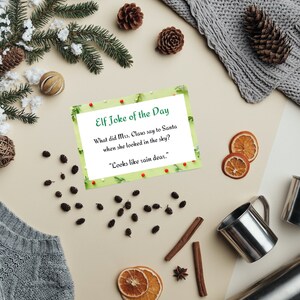 Op de afbeelding: Een feestelijke flat lay met een kerstkaart met de tekst "Elf Joke of the Day", omringd door dennenappels, gedroogde sinaasappelschijfjes, kaneelstokjes en roestvrijstalen bekers. Een grijze trui en besneeuwde dennentakken maken de scène compleet.