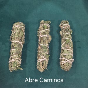 May include: Three bundles of dried green herbs wrapped in white string, arranged vertically. The bundles consist of textured, dried leaves and stems. The text "Abre Caminos" is printed in white at the bottom of the image, centered below the herbs.