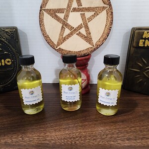 May include: Three small glass bottles with yellow liquid and black caps. Each bottle has a label with the text "Magic Energy 11" and a different name for the oil: "Banishing Oil", "Aceite de Despojo", and "Banishing Oil". The bottles are on a wooden surface with a pentagram symbol in the background.