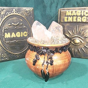 May include: A decorative witch's cauldron with crystals, surrounded by two panels. The panels read "MAGIC" and "MAGIC ENERGY" in gold lettering. The cauldron is brown with black accents and dangling black beads.