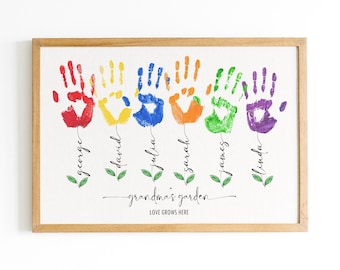 Puede incluir: Una impresión enmarcada con huellas de manos en colores arcoíris formando flores con nombres escritos debajo de cada flor. El texto en la parte inferior dice "Grandma's Garden Love Grows Here".
