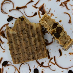 Könnte beinhalten: Zwei Stück Seife mit Kaffeearoma, eine ganz und eine halbiert, auf einer weißen Oberfläche mit braunen Kaffeeresten und Schokoladenspänen.