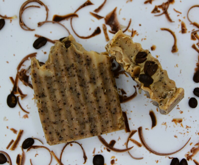 K&ouml;nnte beinhalten: Zwei St&uuml;ck Seife mit Kaffeearoma, eine ganz und eine halbiert, auf einer wei&szlig;en Oberfl&auml;che mit braunen Kaffeeresten und Schokoladensp&auml;nen.