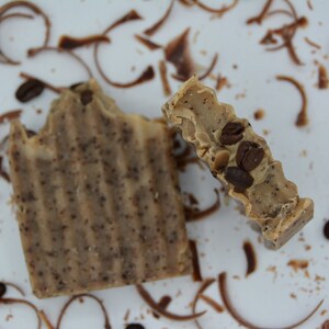 K&ouml;nnte beinhalten: Zwei St&uuml;ck Seife mit Kaffeearoma, eine davon teilweise geschnitten, auf einer wei&szlig;en Oberfl&auml;che mit Kaffeekernen und Schokoladensp&auml;nen. Die Seife ist braun und beige mit eingebetteten Kaffeekernen.