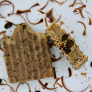 K&ouml;nnte beinhalten: Zwei St&uuml;ck Seife mit Kaffeearoma, eine mit sichtbaren Kaffeebohnen, auf einer wei&szlig;en Oberfl&auml;che mit verstreuten Kaffeepulvern.