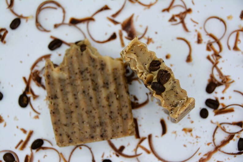 K&ouml;nnte beinhalten: Zwei St&uuml;ck Seife mit Kaffeearoma, eine mit sichtbaren Kaffeebohnen, auf einer wei&szlig;en Oberfl&auml;che mit verstreuten Kaffeepulvern.