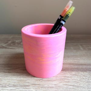 May include: A pink and orange marbled cylindrical pen holder with two markers inside. The markers are black with clear caps and contain orange and yellow ink. The pen holder sits on a wooden surface.