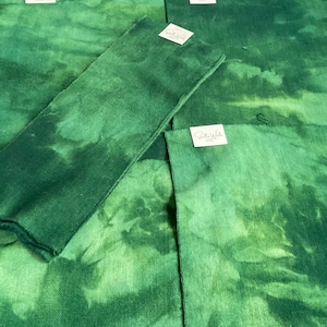 May include: Several pieces of green fabric with a mottled, watercolor-like effect. The fabric has varying shades of green, creating a textured appearance. Small white tags with text are visible on some pieces. The fabric appears to be a craft material.