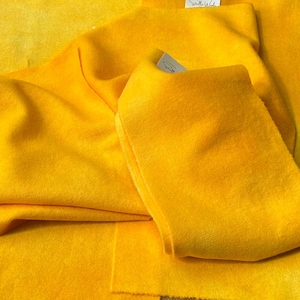 May include: A vibrant yellow fabric with a soft, felted texture, arranged in multiple overlapping layers. Small white rectangular tags with the text Stella Wool are attached to the fabric pieces, which feature a consistent, solid golden-yellow hue.