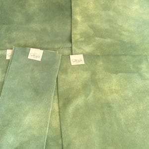 May include: Four pieces of sage green fabric with a mottled, hand-dyed appearance. Each piece features a small white rectangular tag attached to the top edge, printed with the text Stella Wools in a cursive script and smaller block letters.