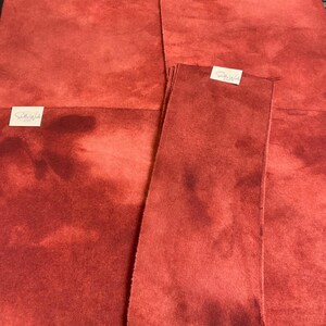 May include: Several pieces of red fabric with a mottled appearance are arranged in a stack. The fabric has a soft, textured surface and a deep red color with darker areas. Small white labels are attached to some pieces.