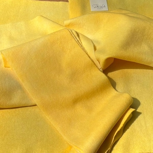 May include: Multiple sheets of bright yellow felt fabric with a soft, fibrous texture. Each piece of material features a small white square label with the text Stella Wools printed in black, attached to the surface of the yellow fabric.