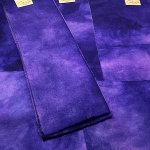 May include: Close-up of several pieces of fabric in varying shades of purple. The fabric has a textured appearance, with darker and lighter areas creating a watercolor effect. Small tags are visible in the upper corners.