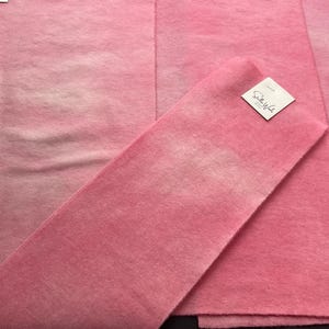May include: Pink fabric with a soft, textured appearance. The fabric is folded and layered, with a tag attached. The color varies from light to a deeper pink. The fabric appears to be a textile material.