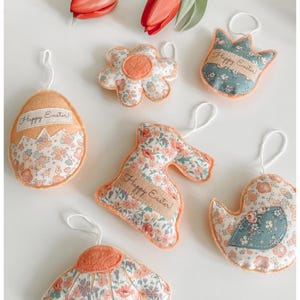 May include: A selection of Easter decorations, including fabric ornaments shaped like an egg, bunny, flower, and bird. The ornaments have a floral pattern with orange details and a hanging string. Some feature the text "Happy Easter!"