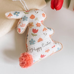 Puede incluir: Un conejito de tela blanca con un patrón floral y una cola de pompón rosa. El conejito tiene una etiqueta que dice "¡Feliz Pascua!"