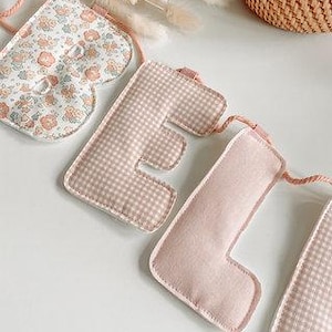 May include: A fabric banner with heart and letter shapes. The heart is patterned with small flowers in peach and white. The letters are in a light pink gingham and solid pink. The banner is strung with a peach-colored cord.