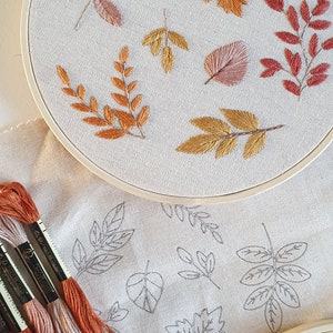 Könnte beinhalten: Nahaufnahme eines unvollendeten Stickprojekts mit verschiedenen Herbstblättern in Orange-, Braun- und Rosatönen. Die Blätter sind auf einem beigefarbenen Stoff gestickt, der in einem hölzernen Stickrahmen gespannt ist. Im Vordergrund sind mehrere Spulen mit Stickgarn in Orange-, Braun- und Rosatönen zu sehen.