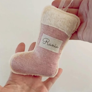 May include: A small, pink Christmas stocking ornament with a white cuff and trim. The name "Rania" is embroidered on a white rectangular patch. A white ribbon is attached for hanging.